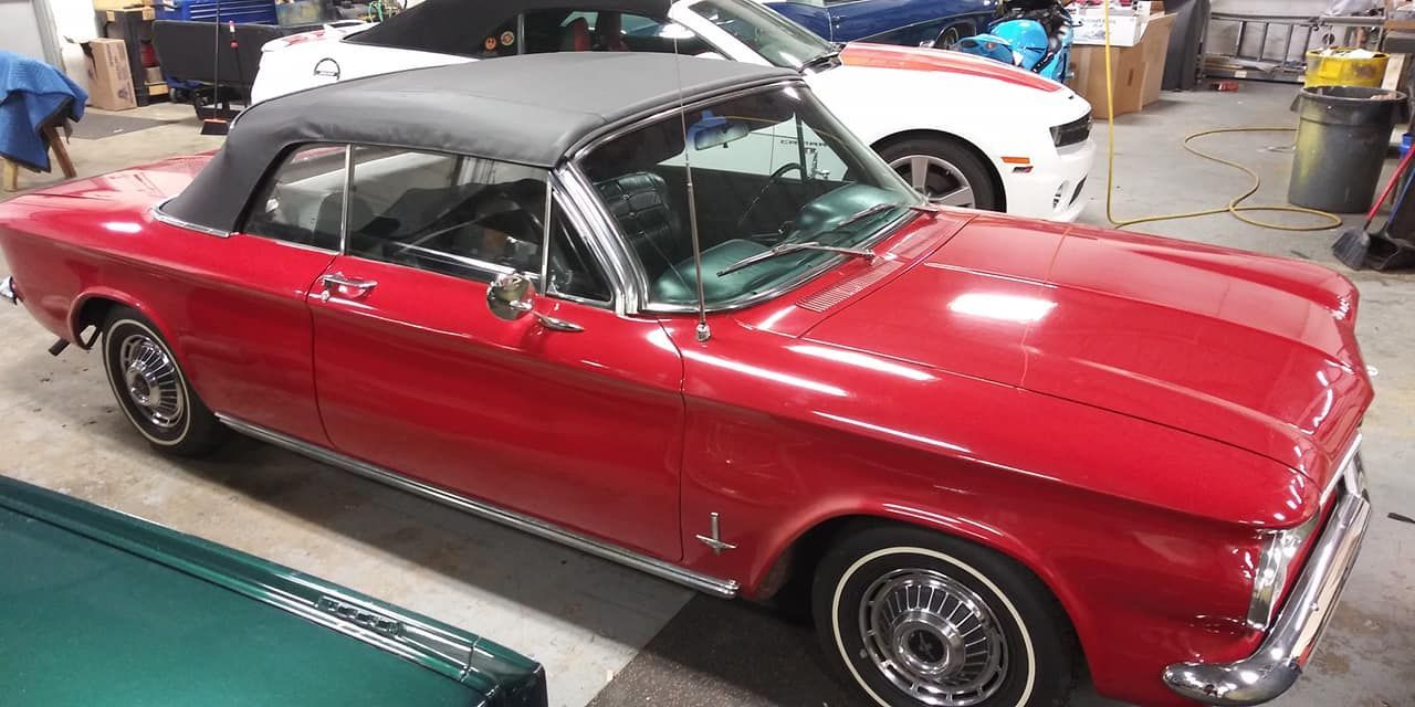 A red vintage convertible with a black soft top parked inside a garage next to a white car.