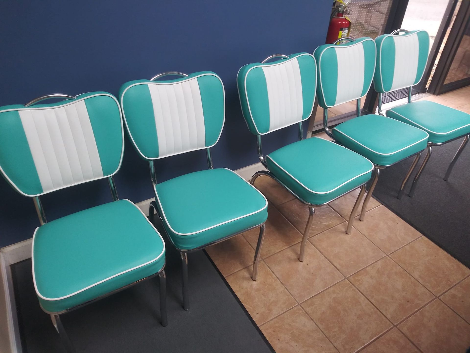 Four matching retro-style diner chairs with teal and white vinyl upholstery arranged in a row against a blue wall.