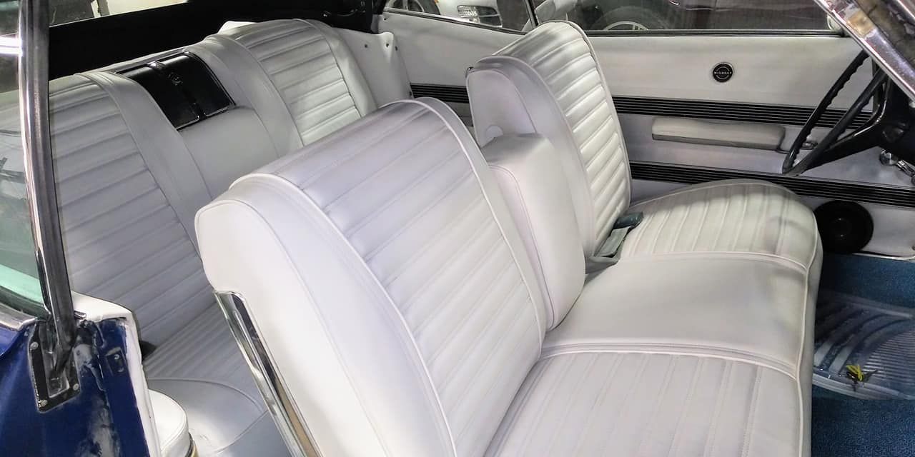 The white vinyl interior of a classic car, featuring a front bench seat with an armrest and a matching back seat.