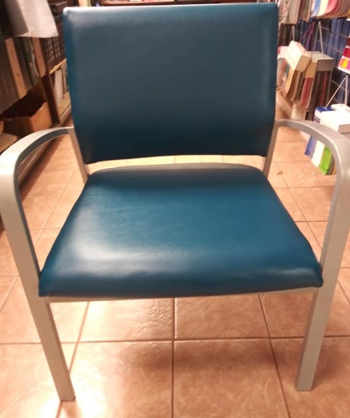 A teal, padded armchair with a silver metal frame, situated on a tiled floor in what appears to be a store.