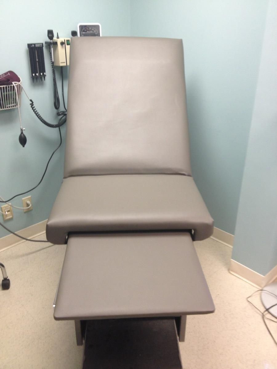 A gray medical examination table with a pull-out footrest in a clinic room, with diagnostic equipment mounted on the wall.