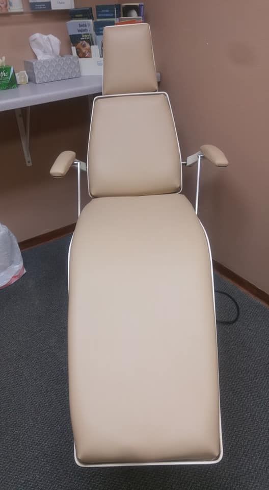 A tan medical or aesthetic procedure chair stands in a room with a desk and supplies in the background.