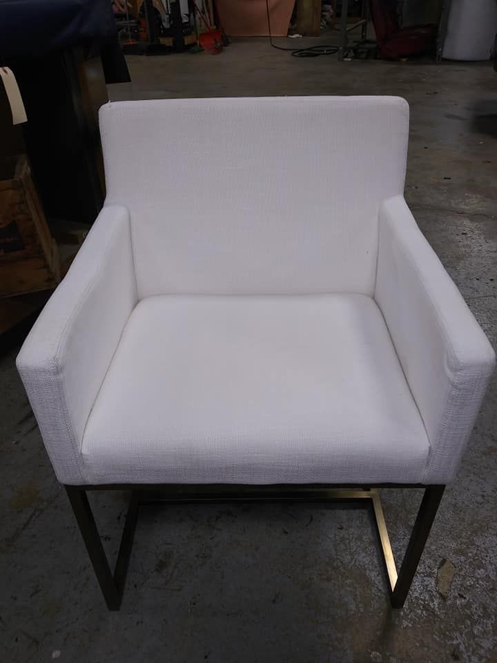 A white upholstered dining chair with armrests and a dark metal, U-shaped base, centered against a workshop background.