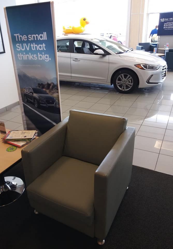 A gray armchair sits in a showroom next to a banner featuring a car, with a white sedan and yellow duck toy in the back.