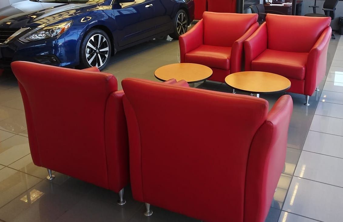 Four red armchairs and two small wooden round tables arranged in a car dealership showroom.