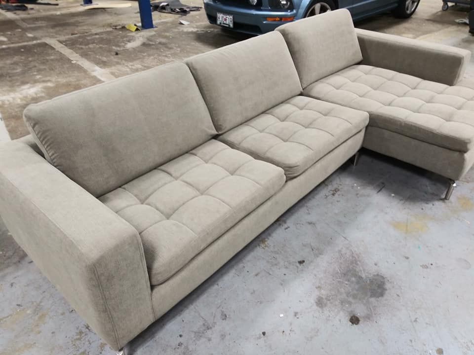 A light beige sectional sofa with tufted seat cushions and metal legs sits in a concrete-floored workshop.