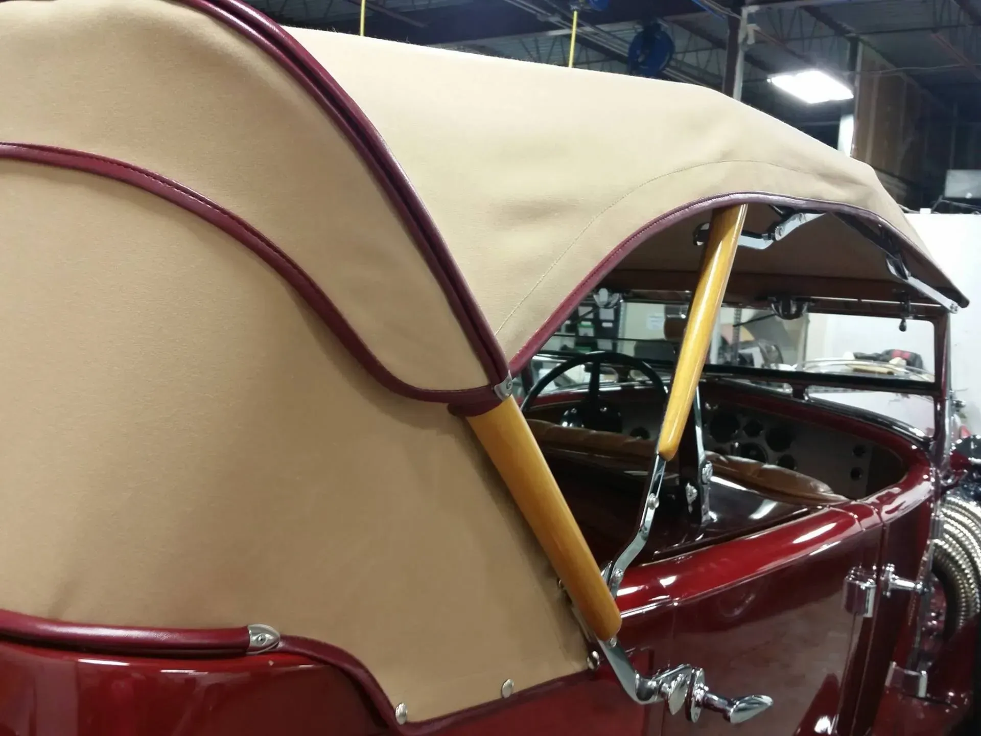 A classic maroon convertible car with a tan fabric soft top roof, viewed from the side in a garage setting.