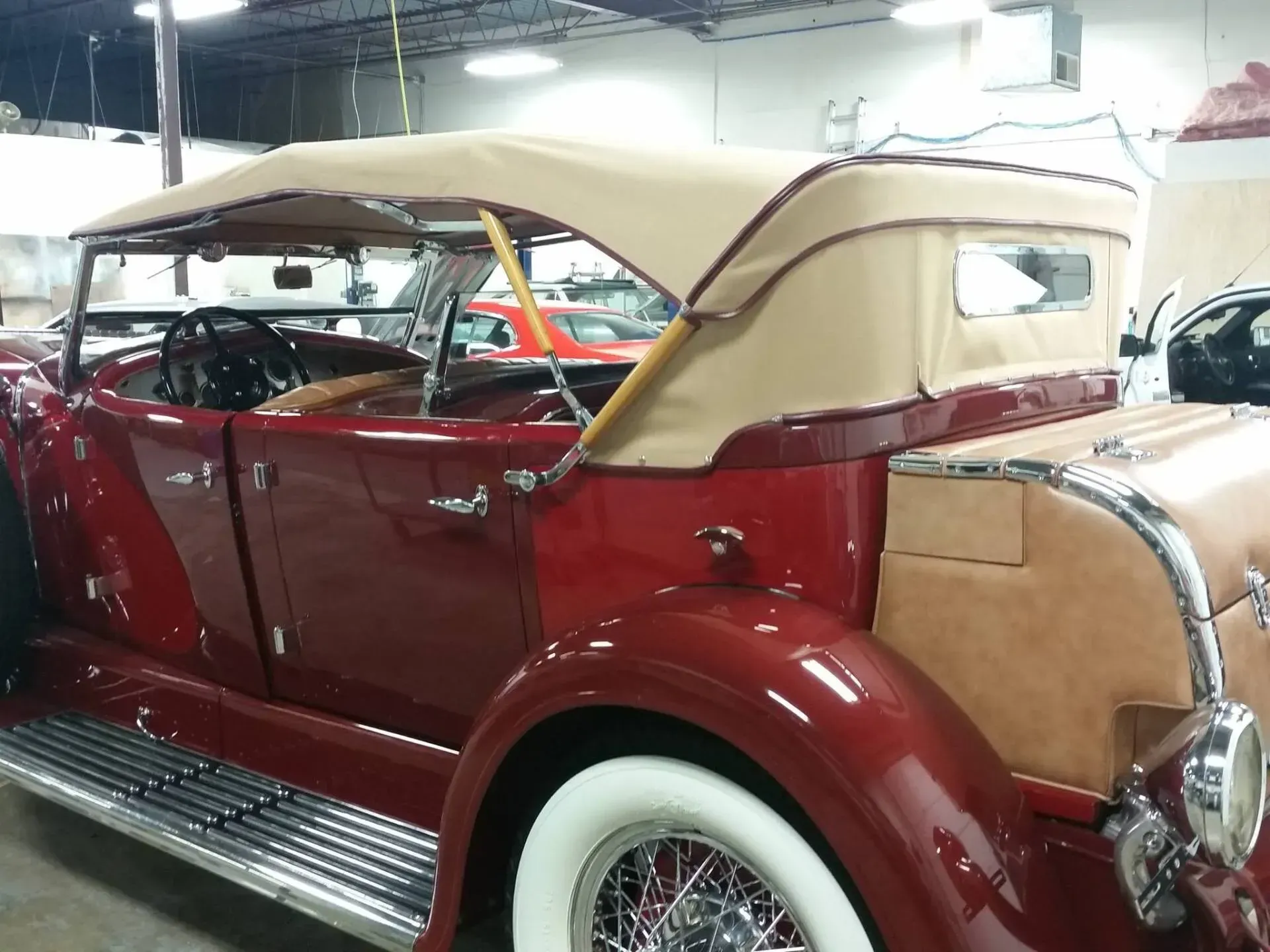 A vintage, deep-red convertible with whitewall tires and a tan soft top parked inside a garage.