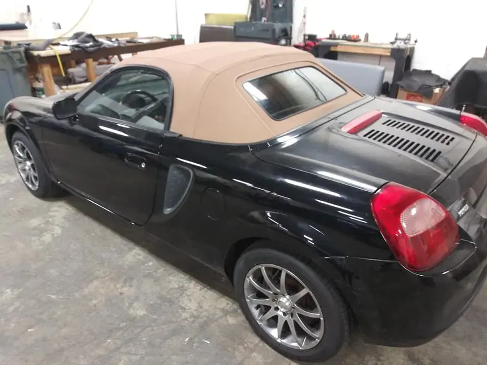 A black Toyota MR2 Spyder with a tan soft-top roof parked in a workshop.