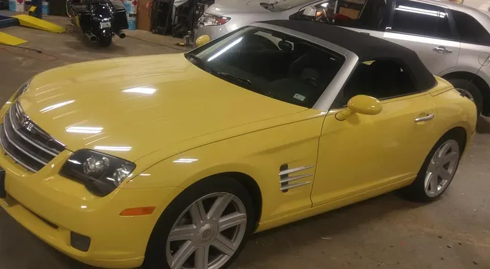 A yellow Chrysler Crossfire convertible parked indoors with a black soft top and silver wheels.