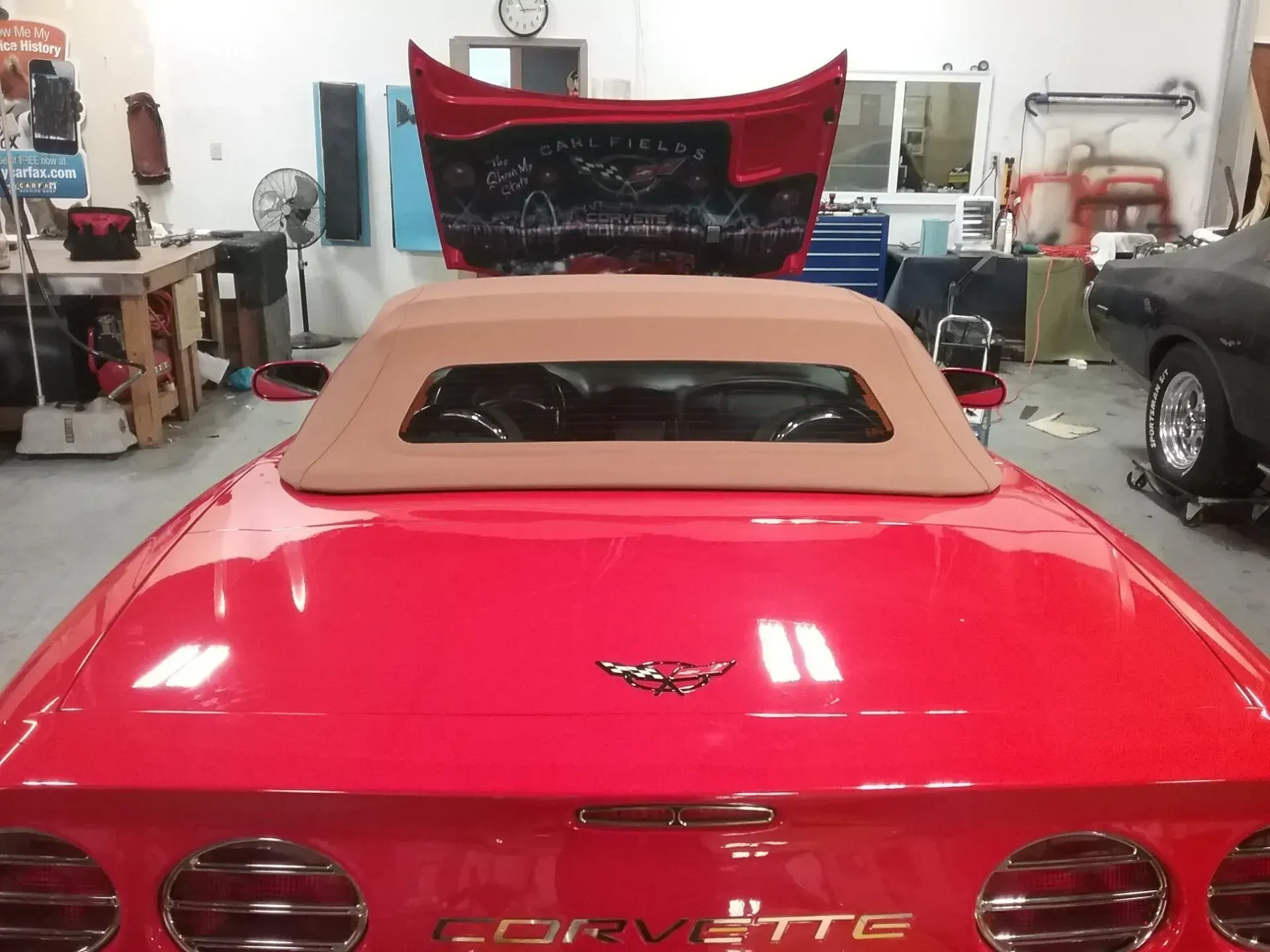 Rear view of a bright red Chevrolet Corvette convertible with the trunk lid open, parked inside an auto repair garage.