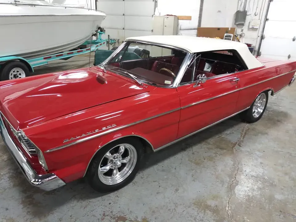 A bright red vintage Ford Fairlane convertible with a white top parked in a garage next to a boat trailer.
