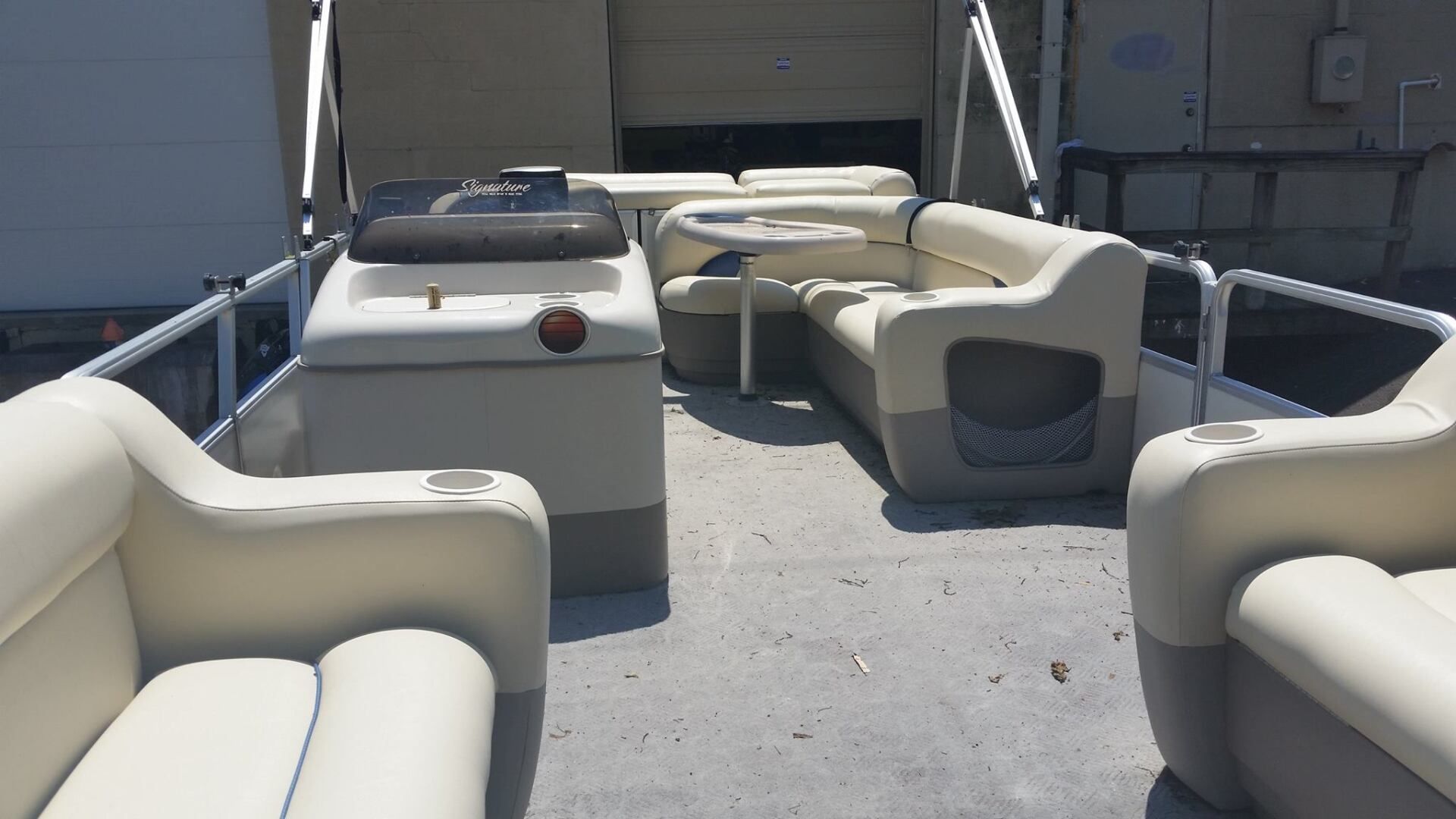 The interior deck of a pontoon boat, featuring beige and gray wrap-around seating, a small table, and a driver's console.
