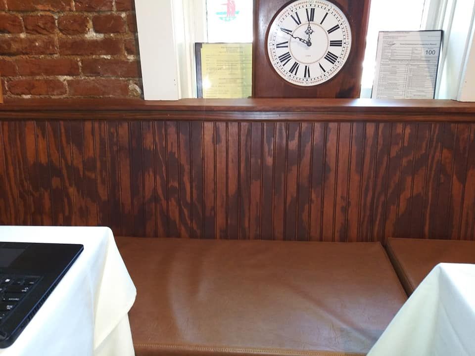 A booth with brown vinyl seating and wood-paneled walls, featuring a large round clock on the windowsill behind it.