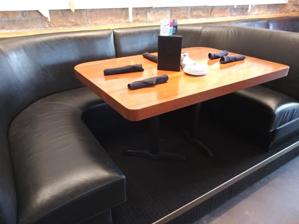 A wooden table with black napkins and a menu holder is set inside a curved, black leather booth in a restaurant.