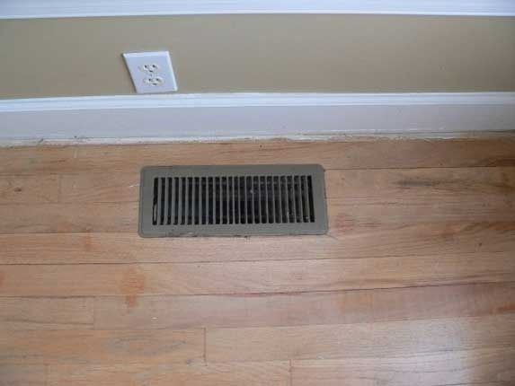 Floor vent in light wood floor, below a white baseboard and tan wall with electrical outlet.