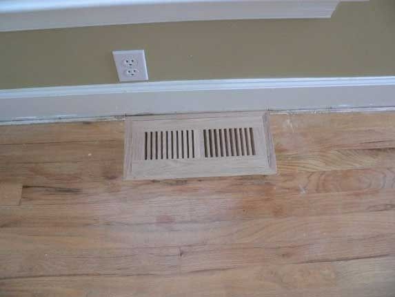 Wooden floor vent with parallel vertical slats on a light brown hardwood floor near a beige wall.