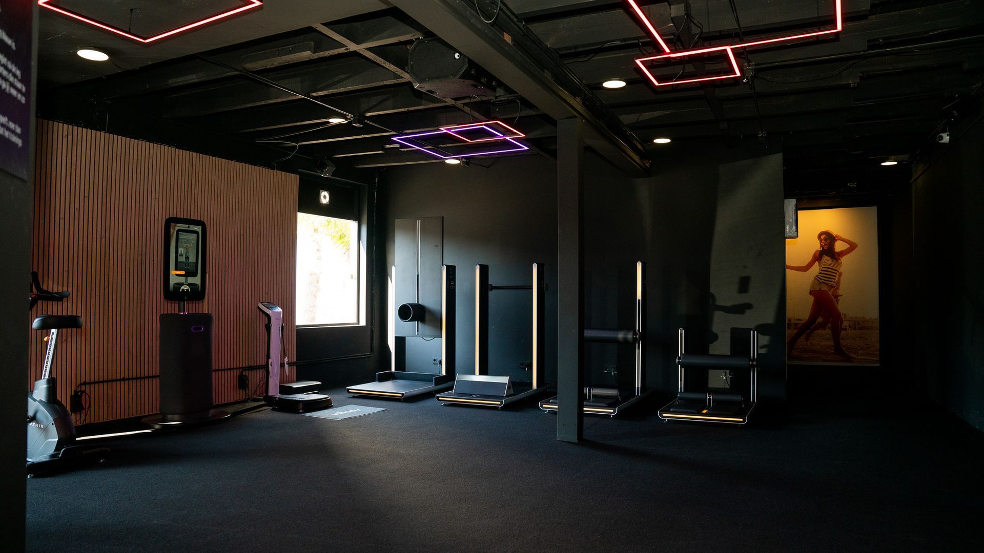 Donkere sportschool met fitnessapparatuur en kunst, verlicht met roze en witte lampen.