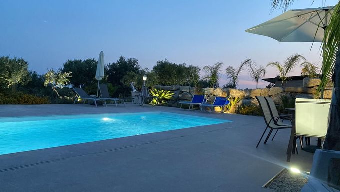 Scena serale a bordo piscina con piscina, lettini, tavolo, sedie e paesaggio illuminato.