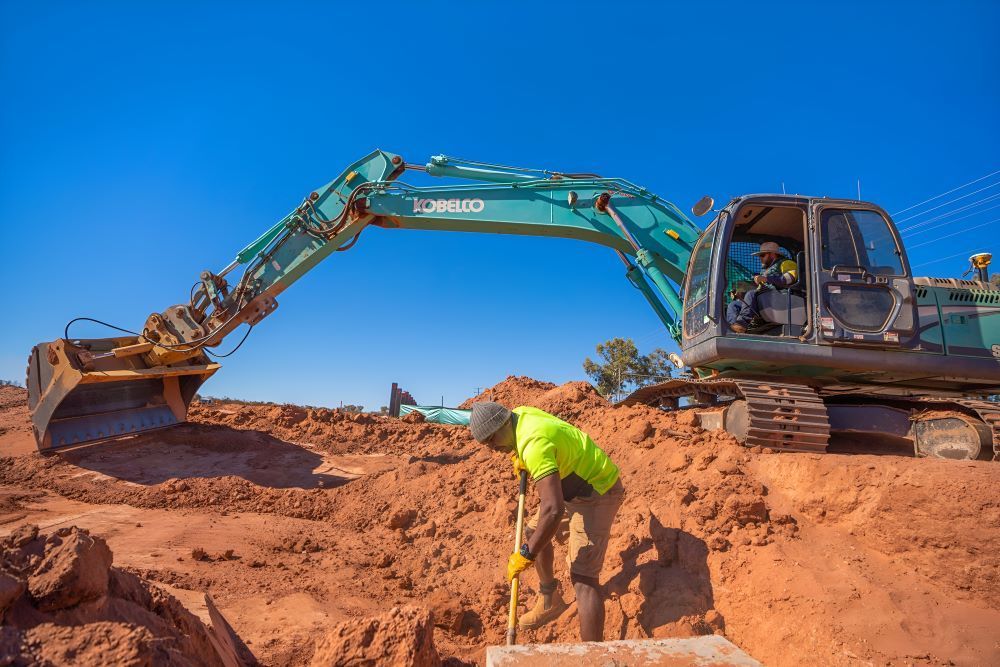A Man is Standing in the Dirt Next to a Large Excavator — ASPLUM & CIVIL In Alice Springs, NT