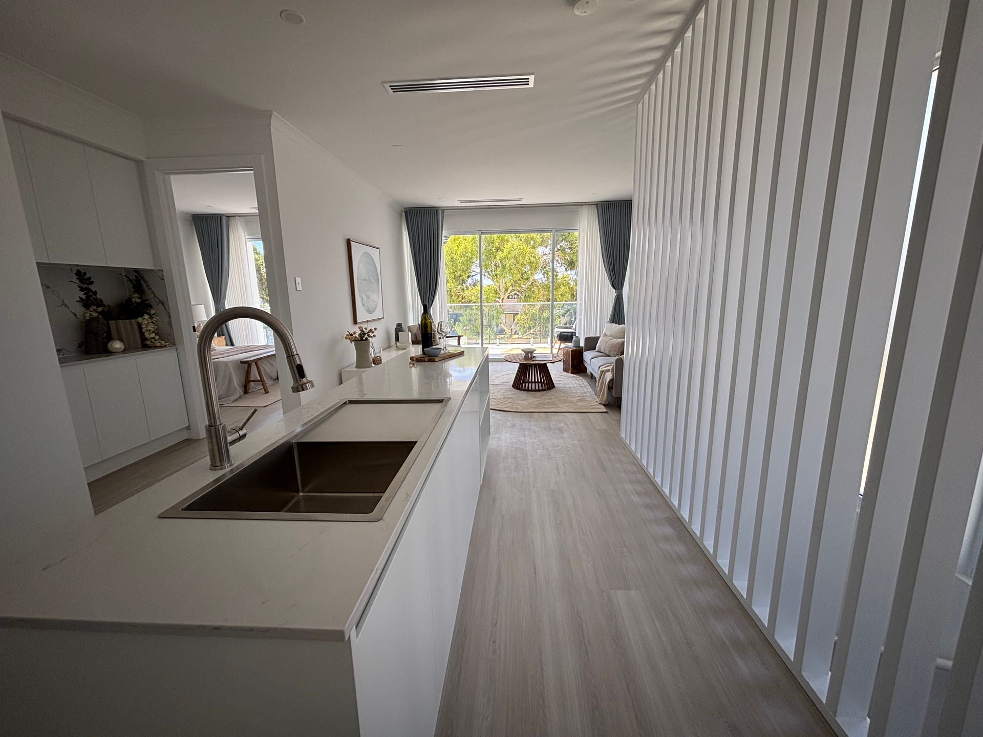 Modern kitchen with long countertop, sink, and view into living area with curtains and light.