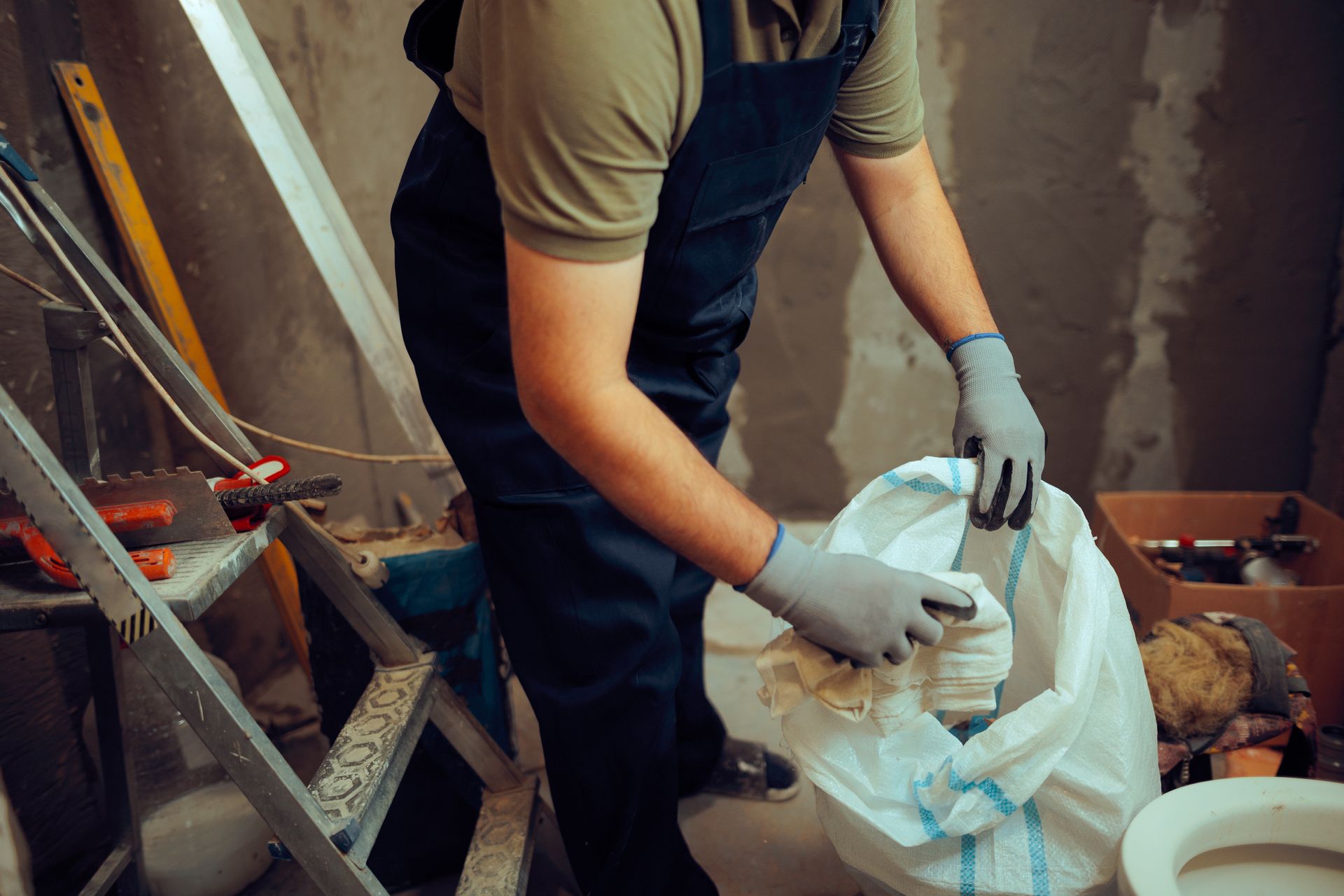 Person in work overalls putting items into a white bag in a construction setting.