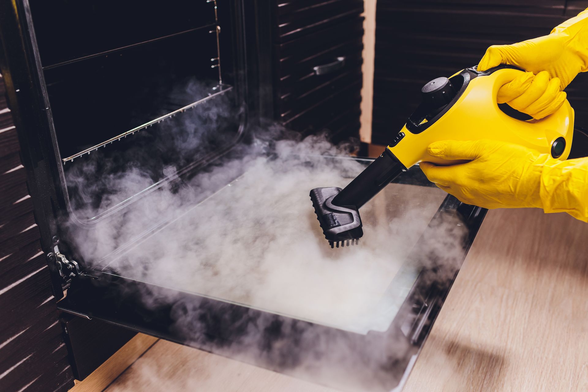Yellow-gloved hands using a steam cleaner inside an oven. Steam fills the oven interior.