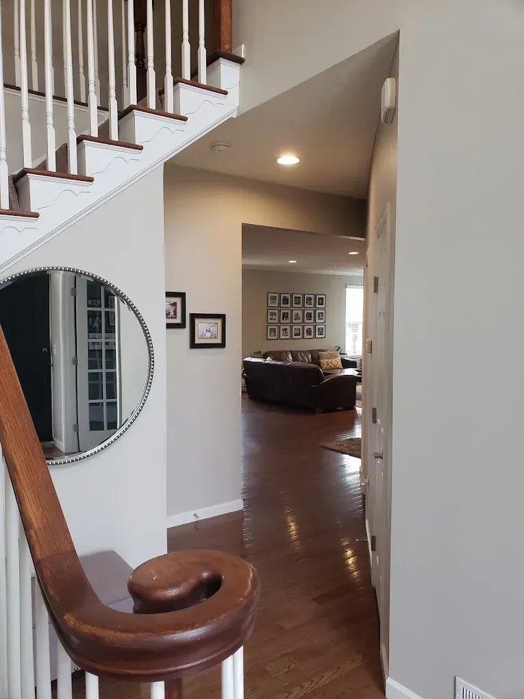A hallway with a wooden floor leads to a living room, viewed from a stairwell. A mirror and framed pictures are visible.