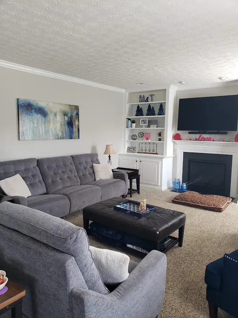 Gray living room with sofa, built-in shelves, fireplace, large TV, and brown leather ottoman.