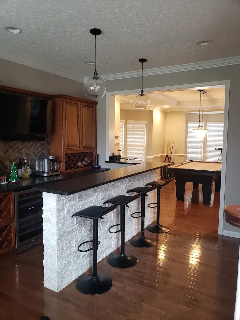 Bar with black countertop, stone facade, stools. Pool table visible in background, pendant lights.