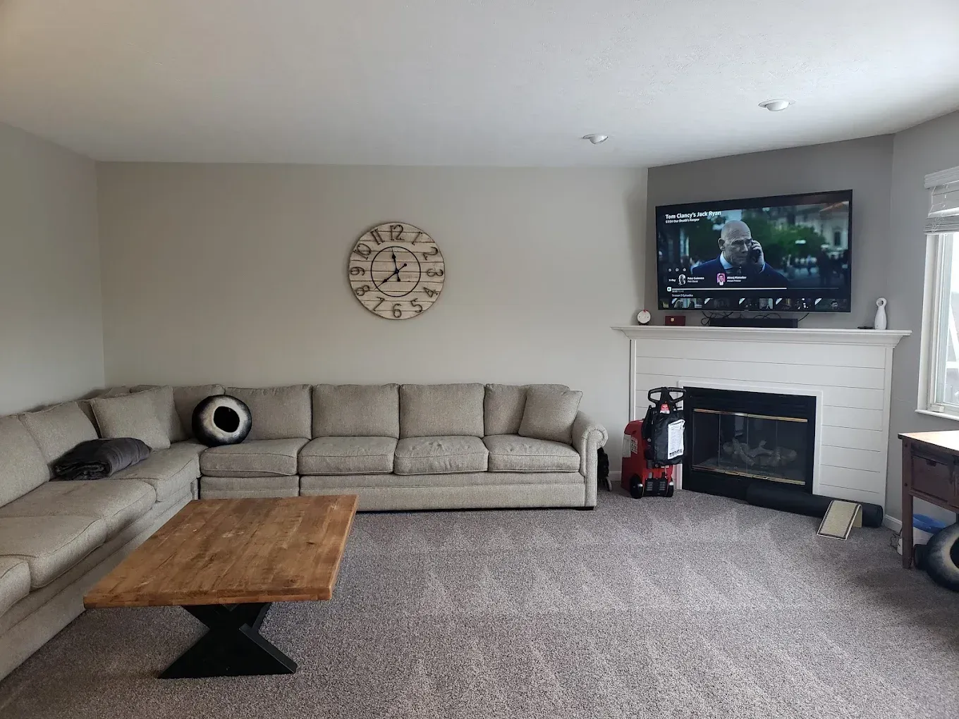 Cozy living room with sectional sofa, fireplace, TV, and large clock. Neutral colors, carpeted floor.