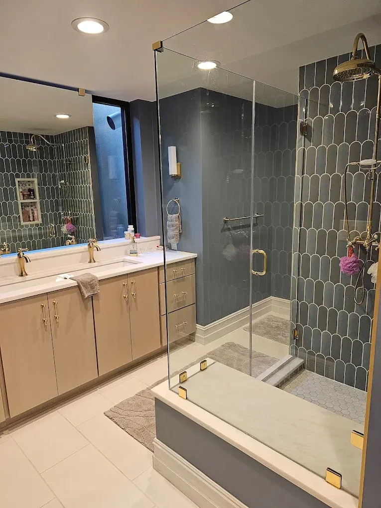 Modern bathroom with glass shower, blue tile accent wall, gold fixtures, and beige cabinets.