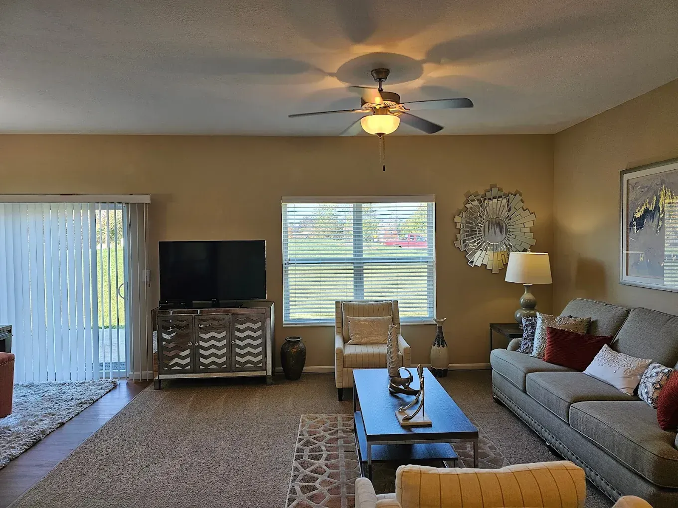 Living room with tan walls, brown rug, TV, sofa, and chairs. Sliding glass door and window provide natural light.