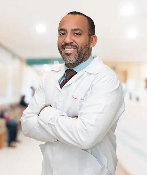 Médico sorridente de jaleco branco, de braços cruzados, em um corredor iluminado de hospital.