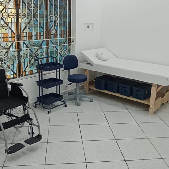Sala de exames vazia com maca, banqueta, andador e caixas organizadoras azuis junto a uma janela gradeada.
