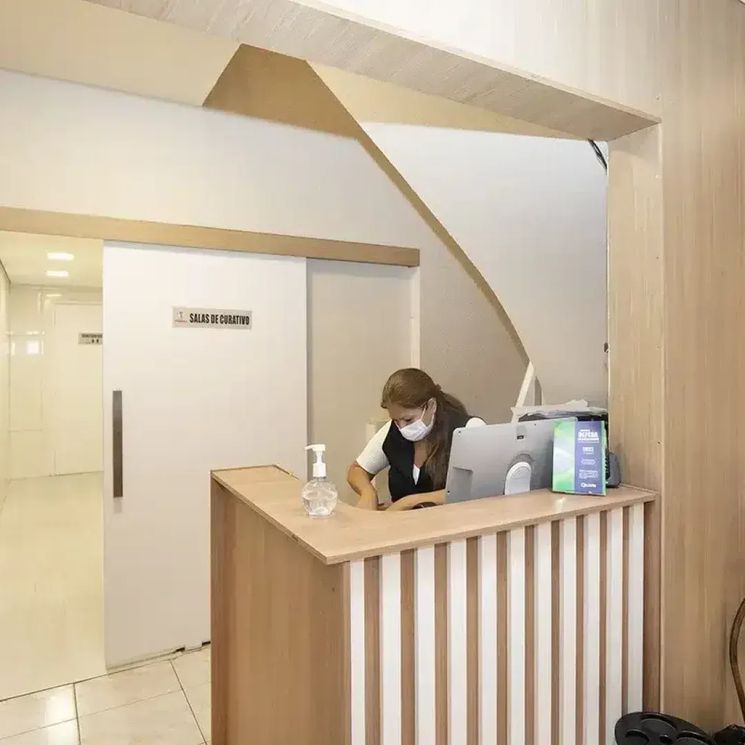 Balcão de recepção em um corredor iluminado, com um funcionário usando máscara trabalhando atrás de um balcão de madeira.