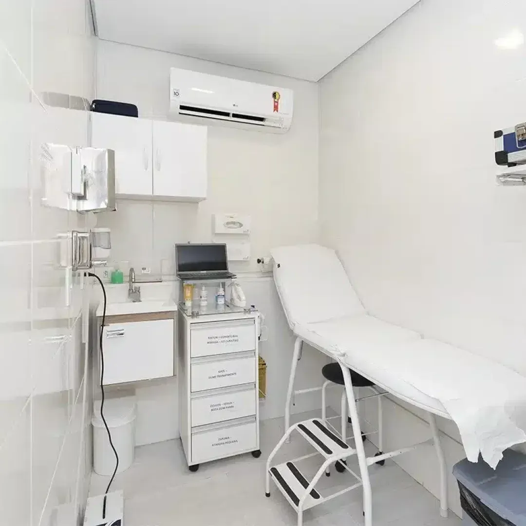 Sala de exames médicos clara e limpa, com maca, armário embutido, pia e ar-condicionado.