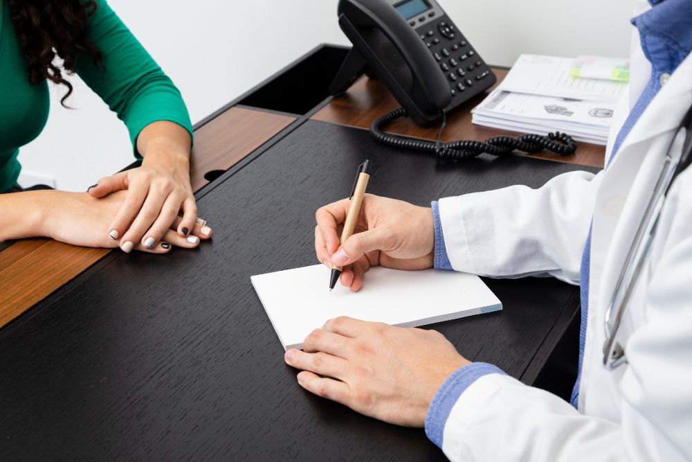 Médico anotando em um bloco de notas em frente a um paciente, sentado à mesa de um consultório.