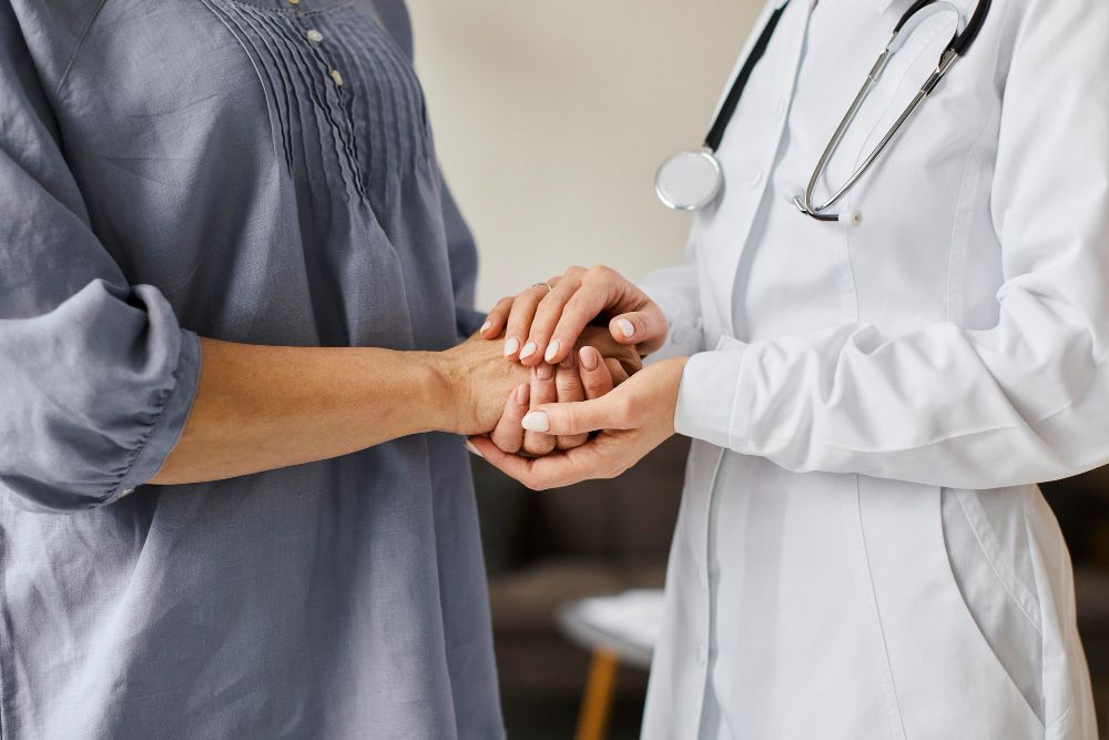 Médico e paciente de mãos dadas em uma clínica, com um estetoscópio visível no jaleco branco do médico.