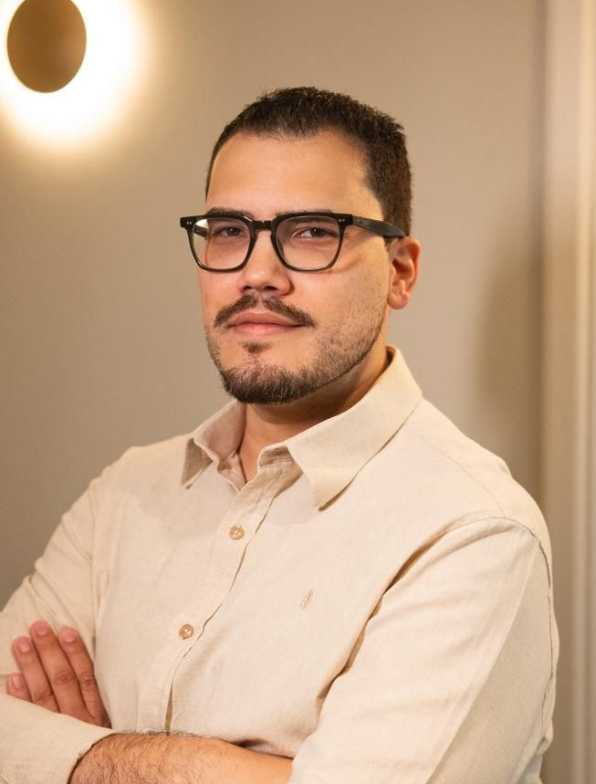 Homem de óculos e braços cruzados, vestindo uma camisa bege, contra um fundo de parede quente em ambiente interno.