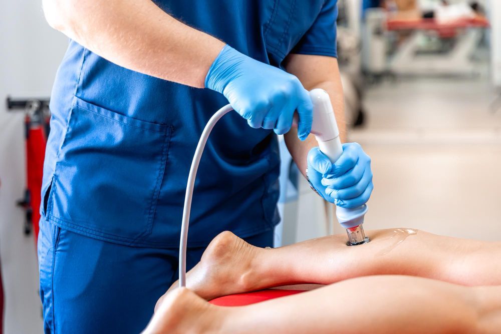 Profissional de saúde com uniforme azul usando um dispositivo de terapia manual na perna de um paciente.