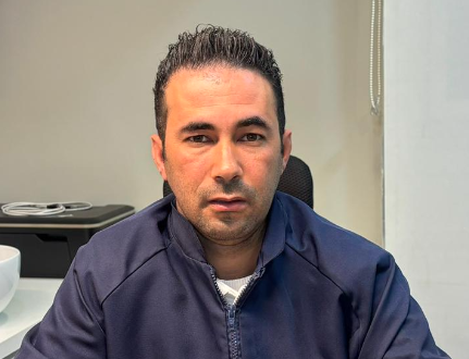 Homem sentado à mesa em um escritório, vestindo um paletó azul-marinho e olhando para a câmera.