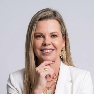 Mulher loira sorridente, vestindo um blazer branco e joias douradas, apoia o queixo na mão contra um fundo claro.