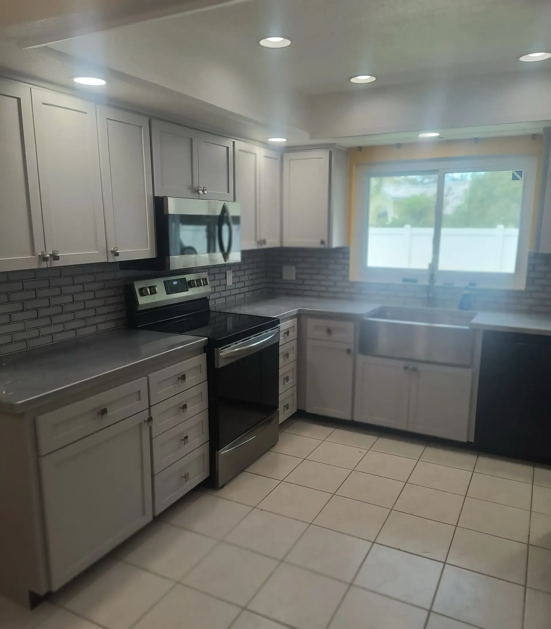 Bright white kitchen with gray cabinets, black stove, tiled floor, and a window over the sink.