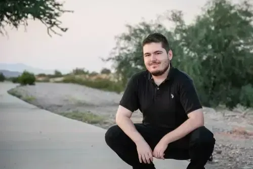 Man crouching on a sidewalk outdoors near a gravel path, wearing a black shirt and smiling at the camera
