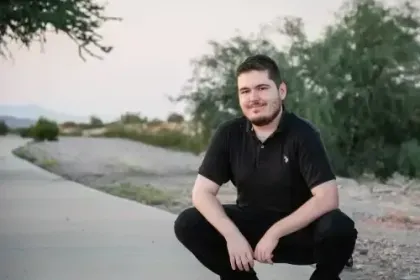 Man crouching on a paved path beside rocks and greenery at dusk, wearing a black shirt.