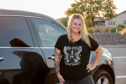Woman standing beside a black SUV outdoors at sunset, wearing a black graphic T-shirt and smiling.