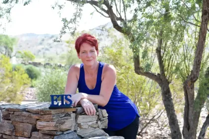 Woman in blue top leaning on stone wall in a sunny outdoor garden