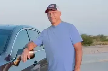 Man standing beside a dark car on a road, leaning on the side mirror, wearing a blue shirt and cap.