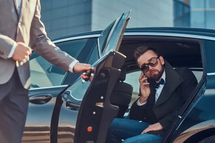 Man in sunglasses talking on phone while seated in a black car as a driver opens the door